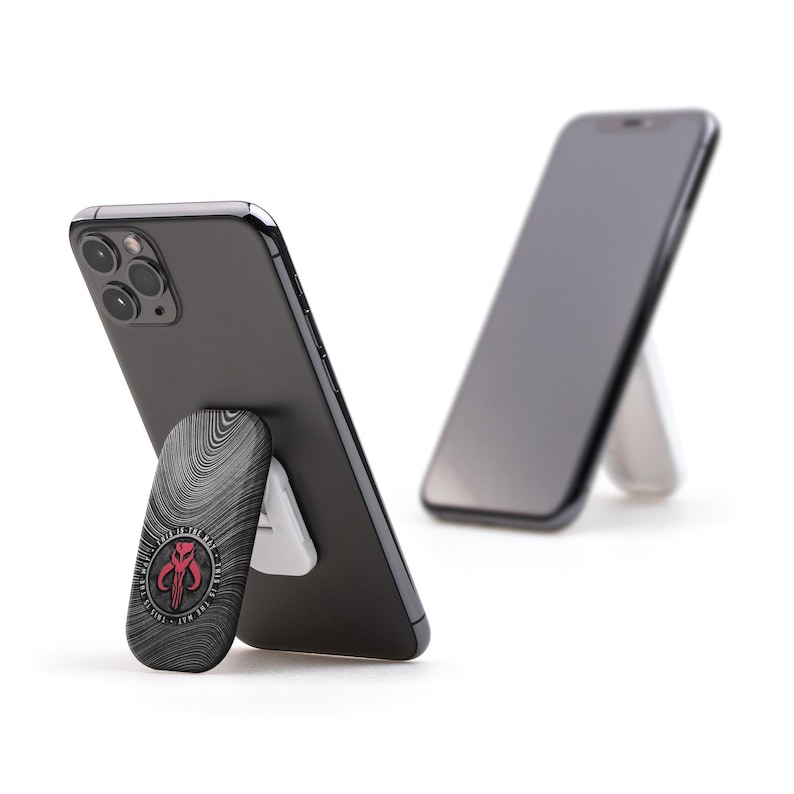 May include: A black smartphone with a textured, gray phone stand featuring a red and white Mandalorian symbol. The phone is propped up in a vertical position.