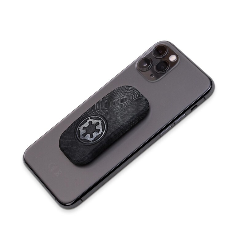 K&ouml;nnte beinhalten: Eine schwarze Handyh&uuml;lle mit Holzmaserung und einem silbernen imperialen Symbol aus Star Wars. Die H&uuml;lle ist an einem schwarzen Smartphone befestigt.