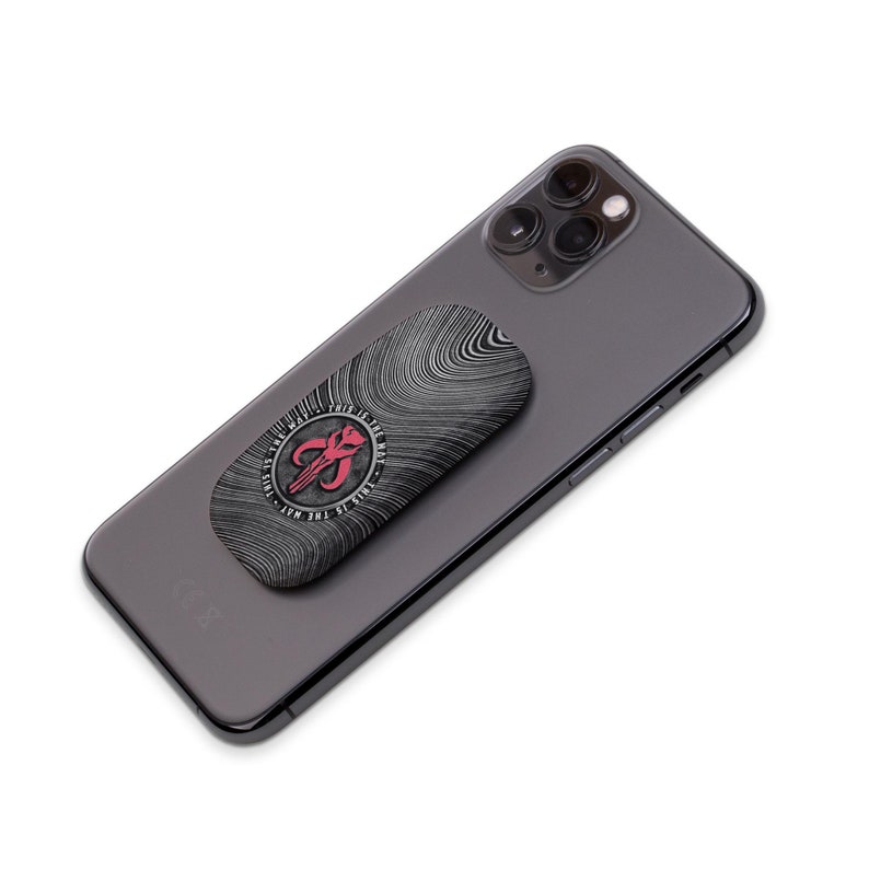 May include: A black and white phone grip with a red Mandalorian symbol and the text "This is the way" in a circle around the symbol. The grip is attached to a black smartphone.