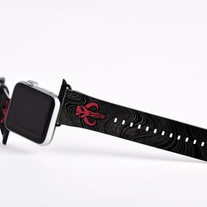 May include: A black and white patterned Apple Watch band with a red Mandalorian symbol. The band is made of silicone and has a black buckle.