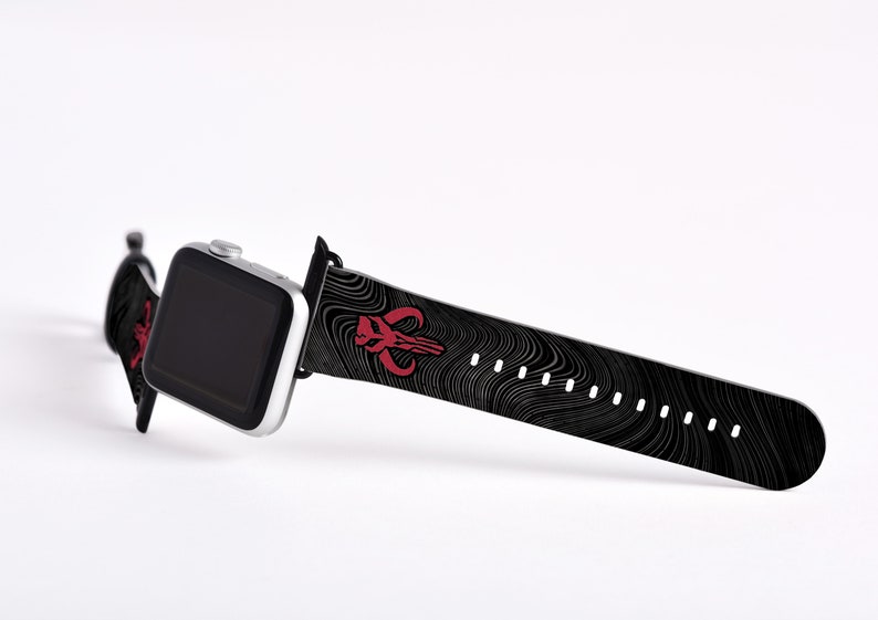 May include: A black and white patterned Apple Watch band with a red Mandalorian symbol. The band is made of silicone and has a black buckle.