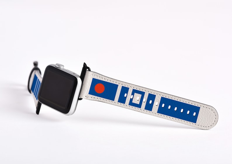 May include: A white Apple Watch band with a blue and red design. The band features a red circle on a blue background, followed by a pattern of blue squares and rectangles.