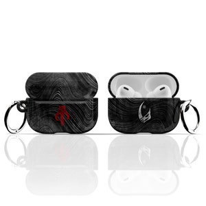 May include: Black and white patterned Airpod case with a red Mandalorian symbol on one side and a white Mandalorian symbol on the other side. The case has a black metal ring attached to it.
