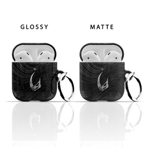 May include: Two black Airpod cases with a white graphic of a horned creature. One case is glossy and the other is matte. Both cases have a silver ring attached.