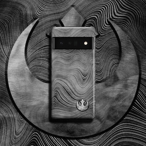 Puede incluir: Una funda de tel&eacute;fono negra con un s&iacute;mbolo de la Alianza Rebelde y un dise&ntilde;o ondulado texturizado. La funda est&aacute; delante de un fondo plateado con un patr&oacute;n ondulado.