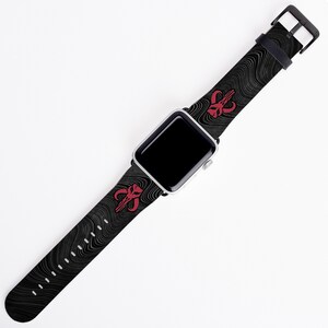 May include: A black and white patterned Apple Watch band with a red Mandalorian symbol. The band is made of a soft, flexible material and is compatible with most Apple Watch models.