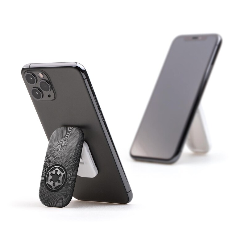 May include: A black smartphone with a black and gray phone grip featuring a wood grain pattern and a black Imperial symbol. The phone is propped up by a white stand, with another phone in the background.