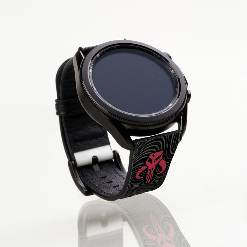 Op de afbeelding: Een zwarte smartwatch met een zwarte band met een rood en zwart Mandalorian-symbool. Het horloge heeft een zwarte wijzerplaat en de band heeft een textuur.