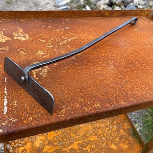 May include: A long, thin, metal tool with a flat, rectangular head and a curved handle. The tool is made of a dark metal and has a rustic appearance. It is lying on a rusty metal surface.