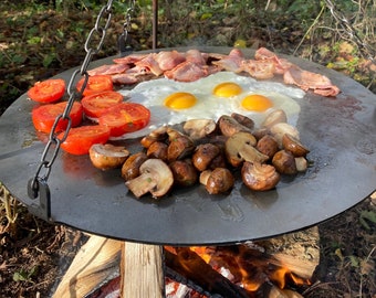 May include: A circular metal griddle, suspended over a wood fire, displaying a cooked breakfast. Sliced tomatoes, fried eggs, bacon rashers, and mushrooms are arranged on the surface. The griddle is suspended by chains.
