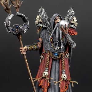 May include: A painted miniature figurine of a wizard holding a skull and a staff with antlers. The wizard is wearing a long, dark grey robe with red accents and a skull-embellished belt. The figurine is standing on a circular base.
