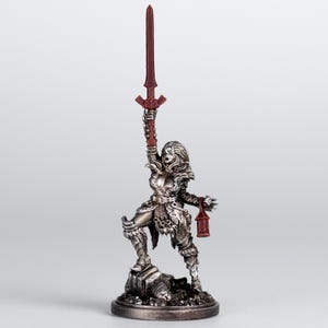 May include: A detailed figurine of a warrior woman, holding a red sword aloft. The figure is silver with intricate armour and stands on a rocky base. A red lantern hangs from her other hand, with a neutral background.