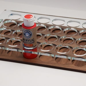 May include: A clear acrylic paint organizer with 24 slots for paint bottles. The organizer is sitting on a wooden base. A red bottle of acrylic craft paint is in one of the slots. The bottle label reads "Crafter's Closet Acrylic Craft Paint Red."