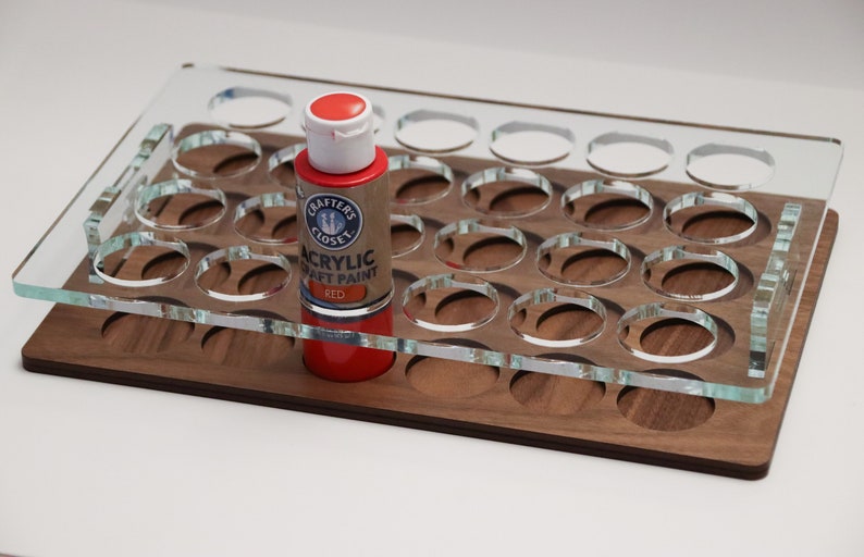 May include: A clear acrylic paint organizer with 24 slots for paint bottles. The organizer is sitting on a wooden base. A red bottle of acrylic craft paint is in one of the slots. The bottle label reads "Crafter's Closet Acrylic Craft Paint Red."