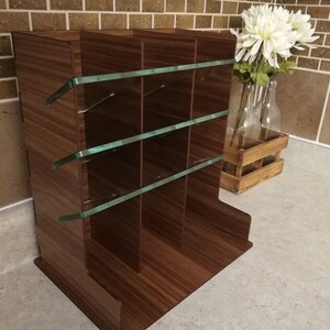 Puede incluir: Un revistero de madera marr&oacute;n con separadores de vidrio. El revistero tiene cuatro compartimentos y est&aacute; sobre una superficie gris claro.