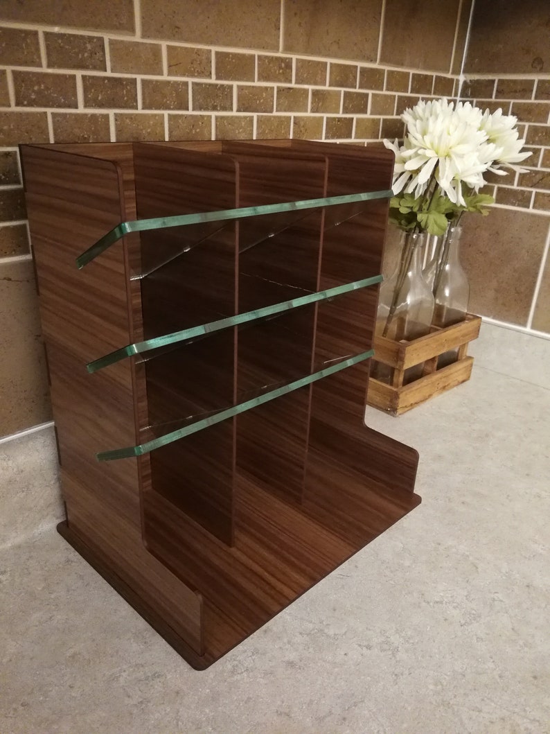 Puede incluir: Un revistero de madera marr&oacute;n con separadores de vidrio. El revistero tiene cuatro compartimentos y est&aacute; sobre una superficie gris claro.