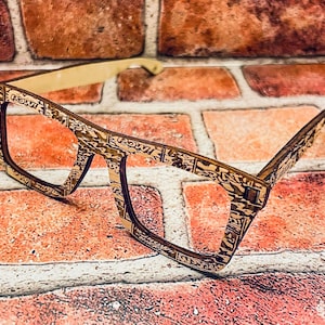 Puede incluir: Un par de gafas de madera con un marco estampado en marr&oacute;n y dorado. Las gafas est&aacute;n apoyadas en una pared de ladrillo.