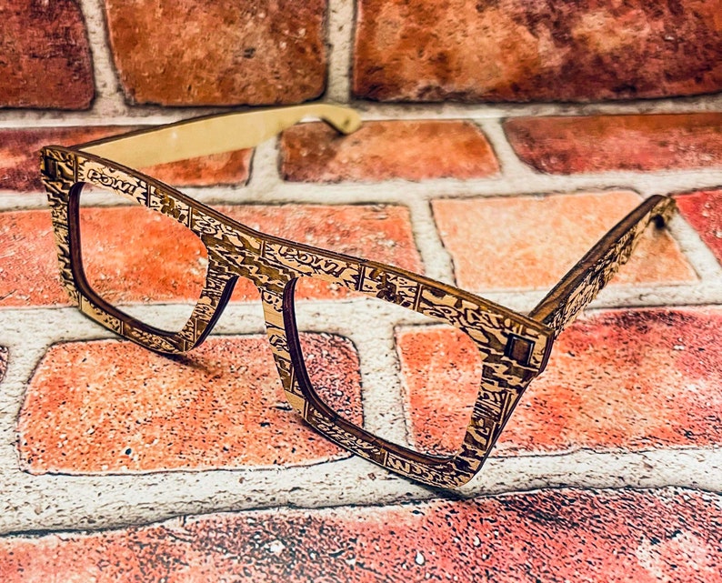 Puede incluir: Un par de gafas de madera con un marco estampado en marr&oacute;n y dorado. Las gafas est&aacute;n apoyadas en una pared de ladrillo.