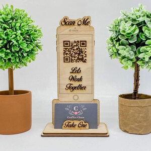 Op de afbeelding: Een houten telefoonstandaard met een QR-code, de tekst "Scan Me", "Let's Work Together" en een visitekaartje met de tekst "Craftee Chaos Take One". De standaard is versierd met twee kleine potplanten.