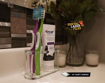 May include: A clear acrylic toothbrush holder with two toothbrushes and a tube of Crest 3D White Whitening Therapy toothpaste. The holder is sitting on a white countertop with two white candles and a vase of greenery in the background.