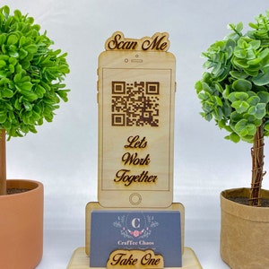 Op de afbeelding: Houten telefoonstandaard met een QR-code en de tekst "Scan Me" en "Let's Work Together". De standaard heeft ook een visitekaartjeshouder met een visitekaartje waarop "CraftTee Chaos" en "Take One" staat.