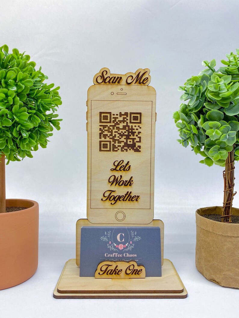 Op de afbeelding: Houten telefoonstandaard met een QR-code en de tekst "Scan Me" en "Let's Work Together". De standaard heeft ook een visitekaartjeshouder met een visitekaartje waarop "CraftTee Chaos" en "Take One" staat.