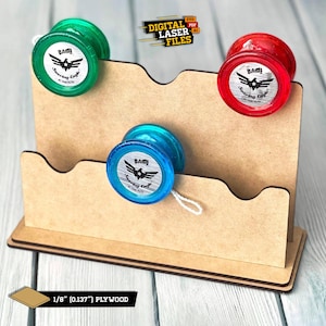 Może przedstawiać: Drewniany stojak z trzema szczelinami do przechowywania jo-jo. Stojak wykonany jest z 3,2 mm sklejki. Na stojaku znajdują się trzy jo-jo, jedno czerwone, jedno niebieskie i jedno zielone. Każde jo-jo ma srebrny wzór z tekstem "BAD!" i "Soaring Eagle" na nim.