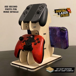 Puede incluir: Archivos de corte láser digital para un soporte de Nintendo Switch. El soporte está hecho de madera contrachapada de 3 mm y acrílico de 6 mm. El soporte puede contener dos mandos, una consola Switch y un cartucho de juego Switch. El soporte se muestra con una consola Switch morada, un mando rojo y negro y una consola Switch negra.