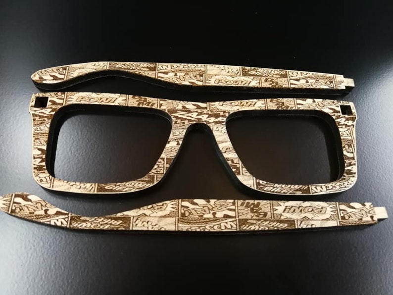 Puede incluir: Un par de gafas de madera con un dise&ntilde;o de impresi&oacute;n de c&oacute;mic. Las gafas est&aacute;n hechas de madera cortada con l&aacute;ser y tienen un acabado marr&oacute;n.