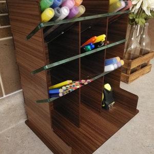 Puede incluir: Un organizador de escritorio de madera con m&uacute;ltiples compartimentos y divisores de vidrio. El organizador est&aacute; lleno de marcadores de varios colores, l&aacute;pices y una cinta m&eacute;trica.