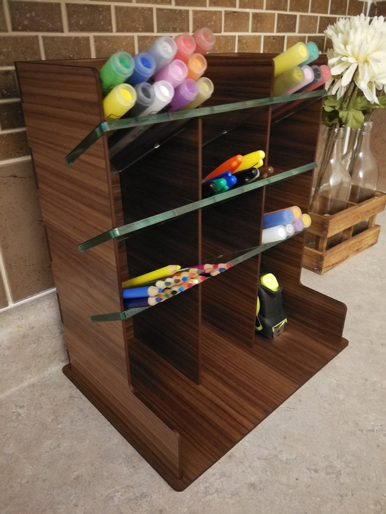 Puede incluir: Un organizador de escritorio de madera con m&uacute;ltiples compartimentos y divisores de vidrio. El organizador est&aacute; lleno de marcadores de varios colores, l&aacute;pices y una cinta m&eacute;trica.