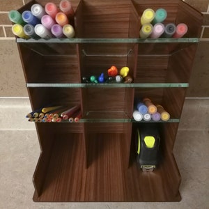 Puede incluir: Un organizador de madera con estantes de vidrio para guardar materiales de arte. El organizador tiene 9 compartimentos y est&aacute; lleno de marcadores de colores, l&aacute;pices y una cinta m&eacute;trica.