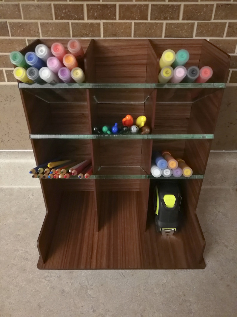 Puede incluir: Un organizador de madera con estantes de vidrio para guardar materiales de arte. El organizador tiene 9 compartimentos y est&aacute; lleno de marcadores de colores, l&aacute;pices y una cinta m&eacute;trica.