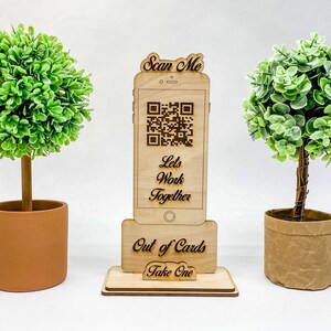 Op de afbeelding: Een houten standaard met een uitsparing in de vorm van een telefoon met een QR-code en de tekst "Scan Me, Let's Work Together, Out of Cards, Take One". Twee kleine gepotte bomen staan aan weerszijden van de standaard.