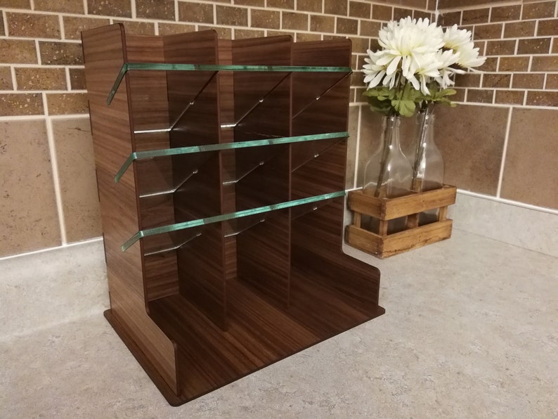 Puede incluir: Un organizador de madera marr&oacute;n con estantes de vidrio y m&uacute;ltiples compartimentos. El organizador est&aacute; sobre una encimera de color gris claro.