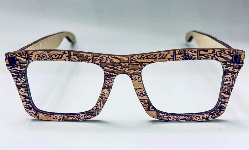 Puede incluir: Un par de gafas con montura de madera marr&oacute;n con un dise&ntilde;o de impresi&oacute;n de c&oacute;mic. Las lentes son transparentes y las monturas tienen una forma cuadrada.