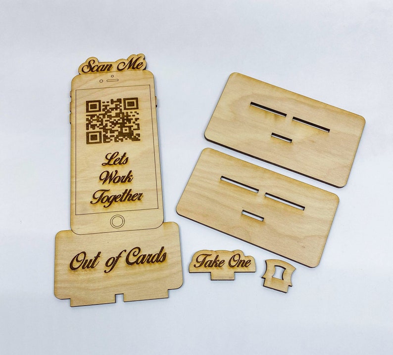 Op de afbeelding: Houten bord met een QR-code, de tekst "Scan Me", "Let's Work Together" en "Out of Cards". Er zijn ook twee houten borden met de tekst "Take One" en een klein houten stuk met een gleuf.