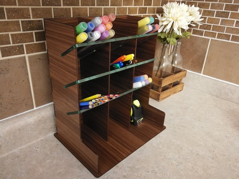 Puede incluir: Un organizador de escritorio de madera con estantes de vidrio. El organizador tiene varios compartimentos para guardar bol&iacute;grafos, l&aacute;pices, marcadores y otros art&iacute;culos de oficina. El organizador es marr&oacute;n con un patr&oacute;n de veta de madera.