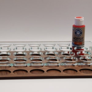 May include: A clear acrylic paint organizer with 24 round slots, sitting on a wooden base. A red bottle of acrylic craft paint is on top of the organizer. The bottle label reads "Crafter's Closet Acrylic Craft Paint Red."