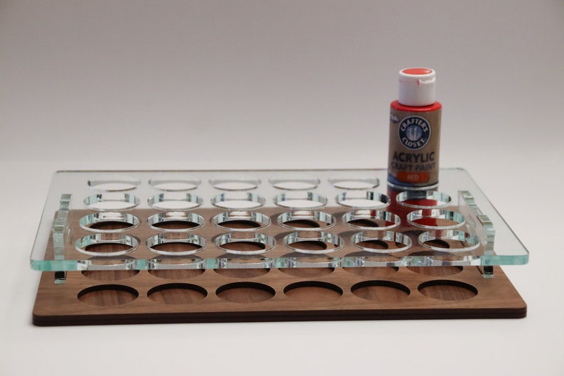 May include: A clear acrylic paint organizer with 24 round slots, sitting on a wooden base. A red bottle of acrylic craft paint is on top of the organizer. The bottle label reads "Crafter's Closet Acrylic Craft Paint Red."