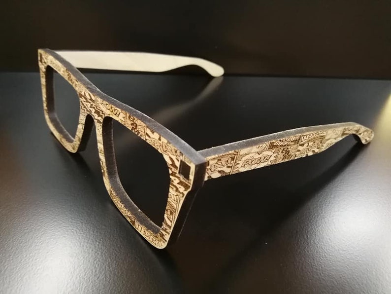 Puede incluir: Un par de gafas de madera marr&oacute;n con un patr&oacute;n de c&oacute;mic. Las gafas tienen un puente nasal de pl&aacute;stico blanco.