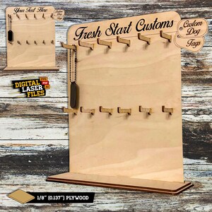 Op de afbeelding: Een houten displaystandaard voor gepersonaliseerde hondenpenningen. De standaard heeft een bord met de tekst "Fresh Start Customs" en heeft haken om penningen aan te hangen. De standaard is gemaakt van 1/8 inch multiplex.
