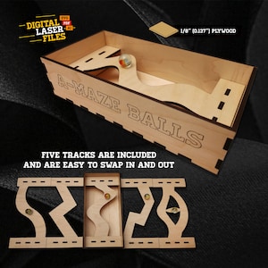 May include: A wooden A-Maze Balls game with five interchangeable tracks. The box and tracks are made of light-colored plywood. The text "A-MAZE BALLS" is engraved on the box. The image also includes the text "FIVE TRACKS ARE INCLUDED AND ARE EASY TO SWAP IN AND OUT."