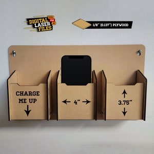 Może przedstawiać: Drewniana stacja ładowania telefonu w kolorze brązowym z trzema przegródkami. Środkowa przegródka ma 4 cale szerokości i może pomieścić telefon. Lewa przegródka zawiera napis "CHARGE ME UP" i strzałkę skierowaną w dół. Prawa przegródka ma wymiar 3,75 cala i strzałkę skierowaną w górę. Stacja jest wykonana z 1/8 cala sklejki.