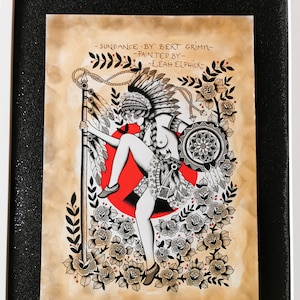 Op de afbeelding: Een zwart-witte illustratie van een indiaanse vrouw met een hoofdtooi die een speer vasthoudt. Ze is omringd door rozen en de tekst "SUNDANCE BY BERT GRIMM - PAINTED BY LEAH ELPHICK".