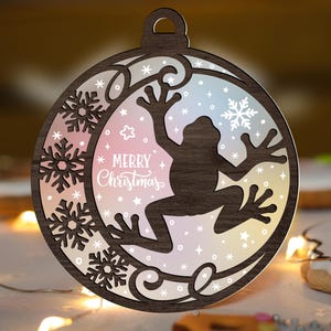 May include: Wooden Christmas ornament with a frog silhouette inside a circular frame. The frame features snowflakes and the words "Merry Christmas." The background has a gradient of pink, blue, and yellow.