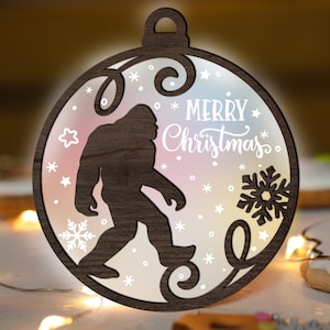 May include: A wooden Christmas bauble shaped like a circle with a silhouette of Bigfoot in the centre. The bauble has a white background with snowflakes and stars. The text "Merry Christmas" is written in white cursive lettering.