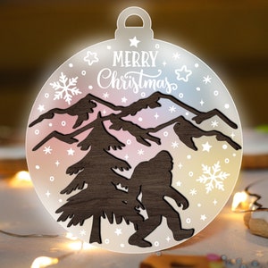 May include: A white, round Christmas bauble with a brown silhouette of a mountain range, a pine tree, and a Bigfoot. The bauble has a white snowflake, stars, and the text "Merry Christmas" on it.