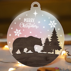 May include: A wooden Christmas ornament shaped like a bauble with a brown bear silhouette, two pine trees, and the text "Merry Christmas" in white lettering on a pink and blue ombre background.