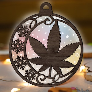 May include: A wooden Christmas ornament shaped like a bauble with a cannabis leaf design. The ornament is decorated with snowflakes and stars.